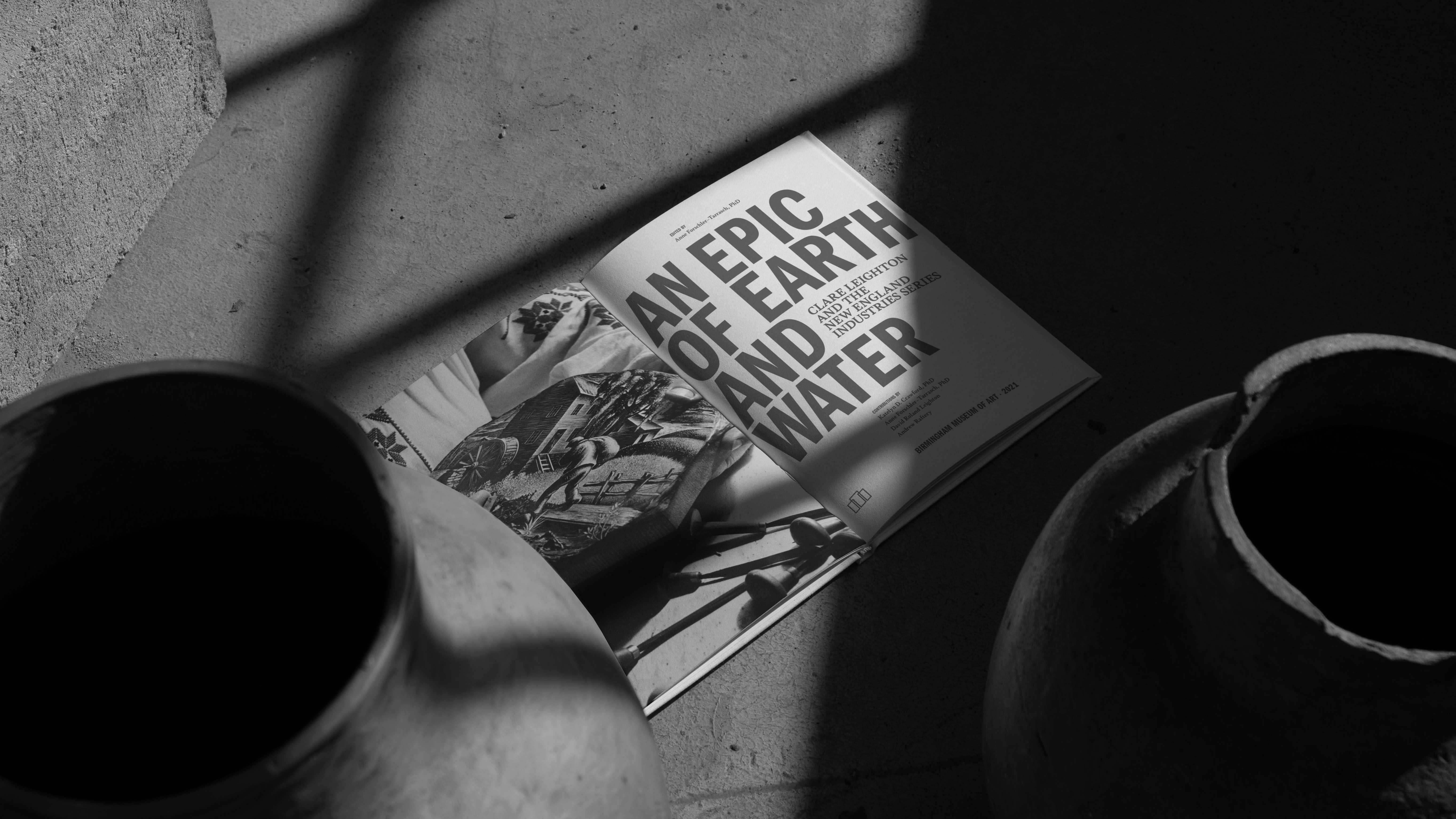 black and white image of an open book sitting on the floor surrounded by clay pots harsh light and shadows